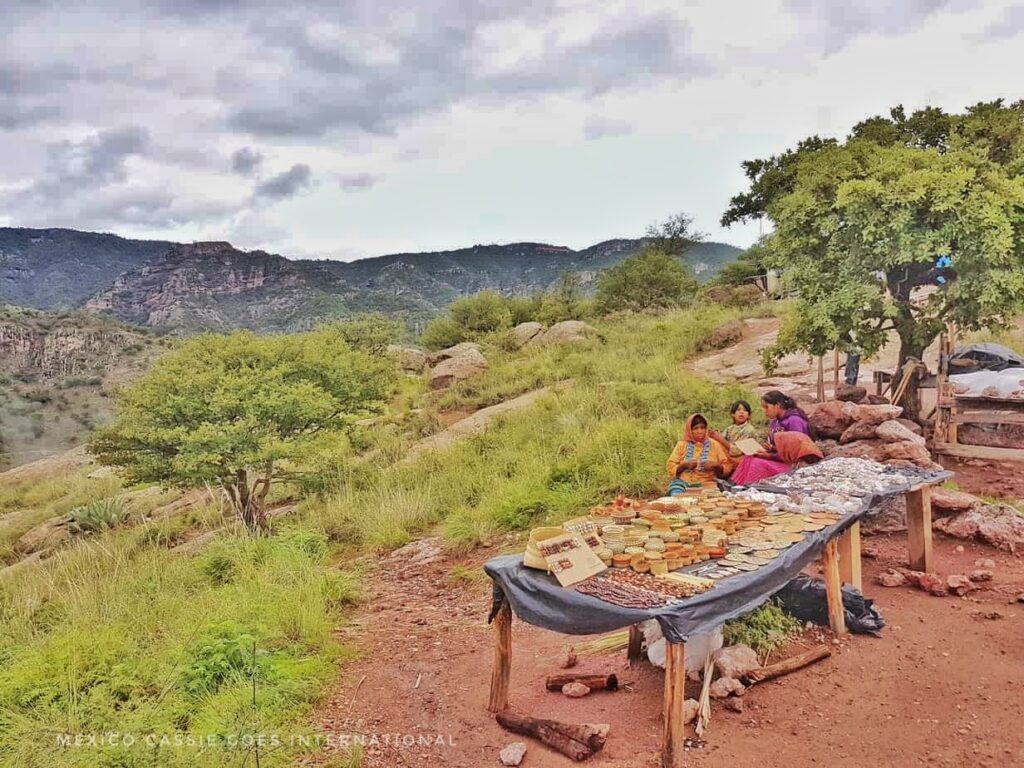 table with wooden items for sale. three women siting by it, hills and greenery in background