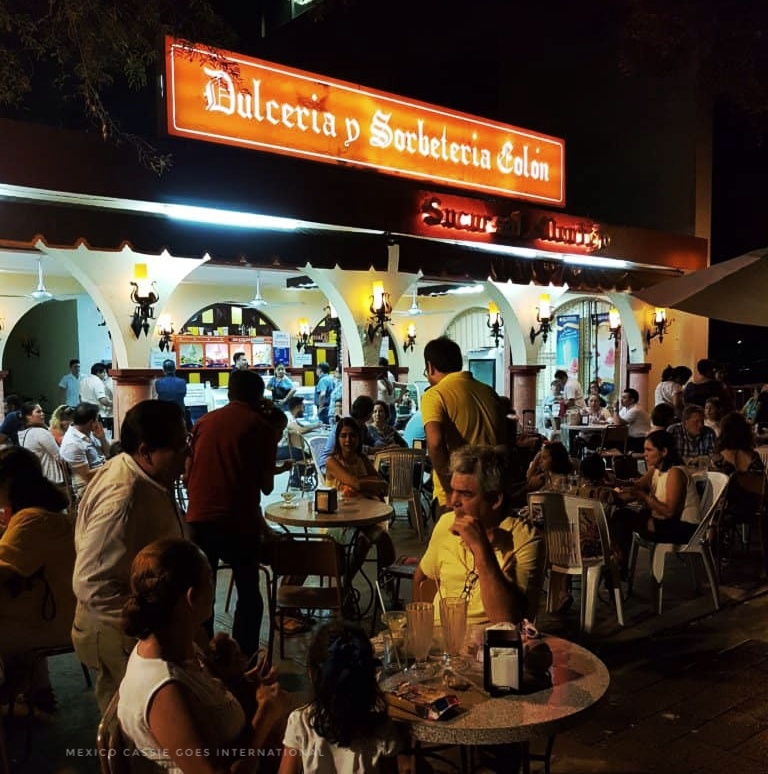 night shot of people at tables under an orange sign reading Dulceria y Sobeteria Colon