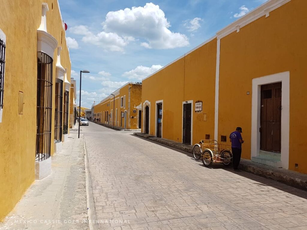 empty street. yellow buildings. one man with bike standing at side.