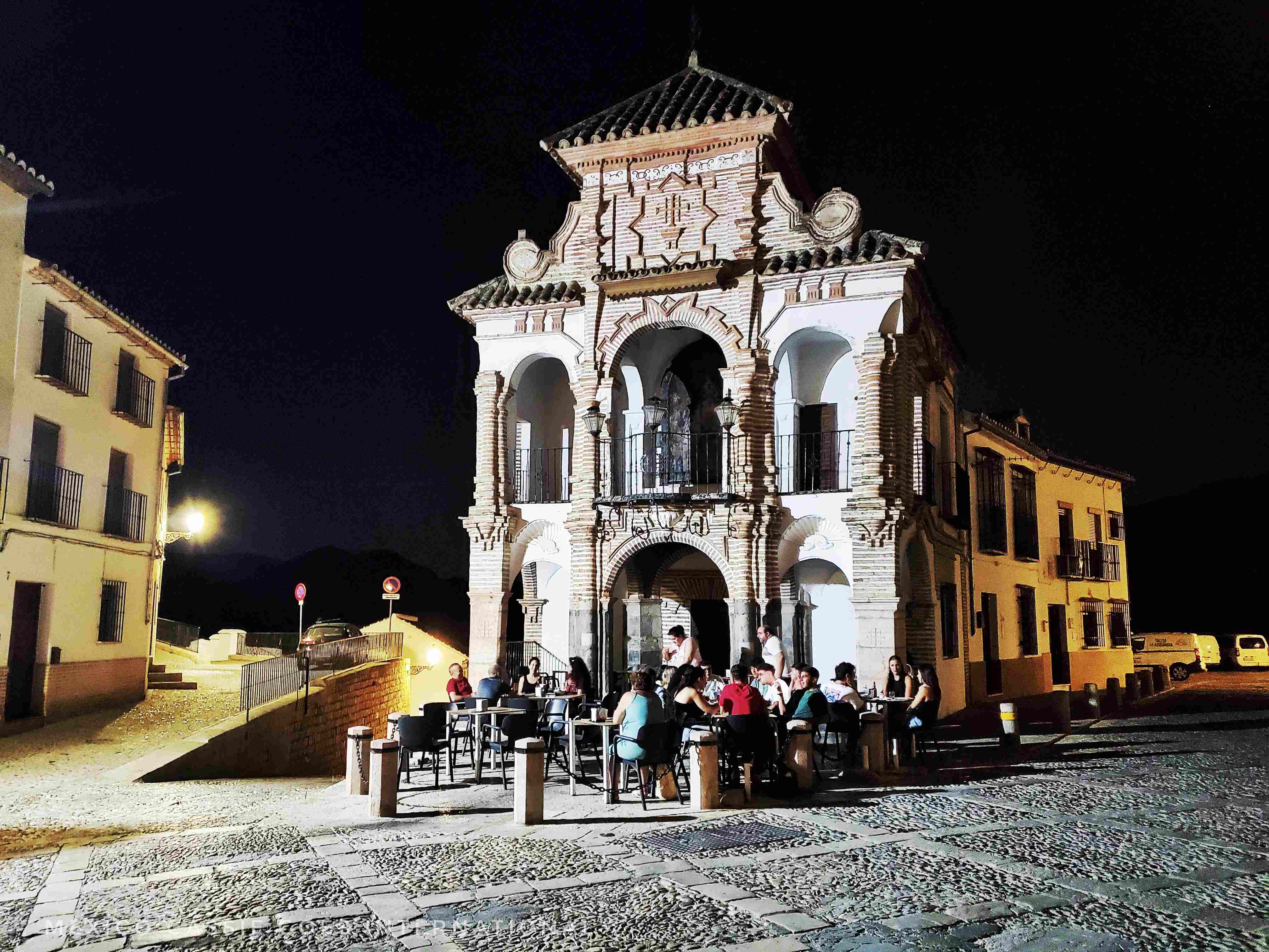 view of a white facade chapel at night. people drinking at tables under the arches