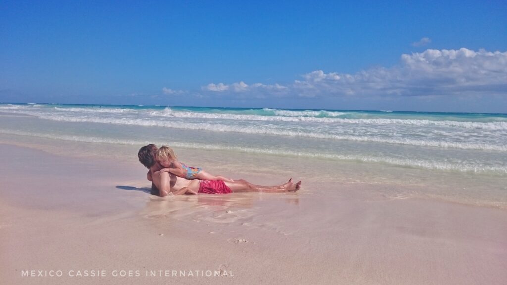 man in red shorts lying on beach with small child on top of him . Perfect blue water