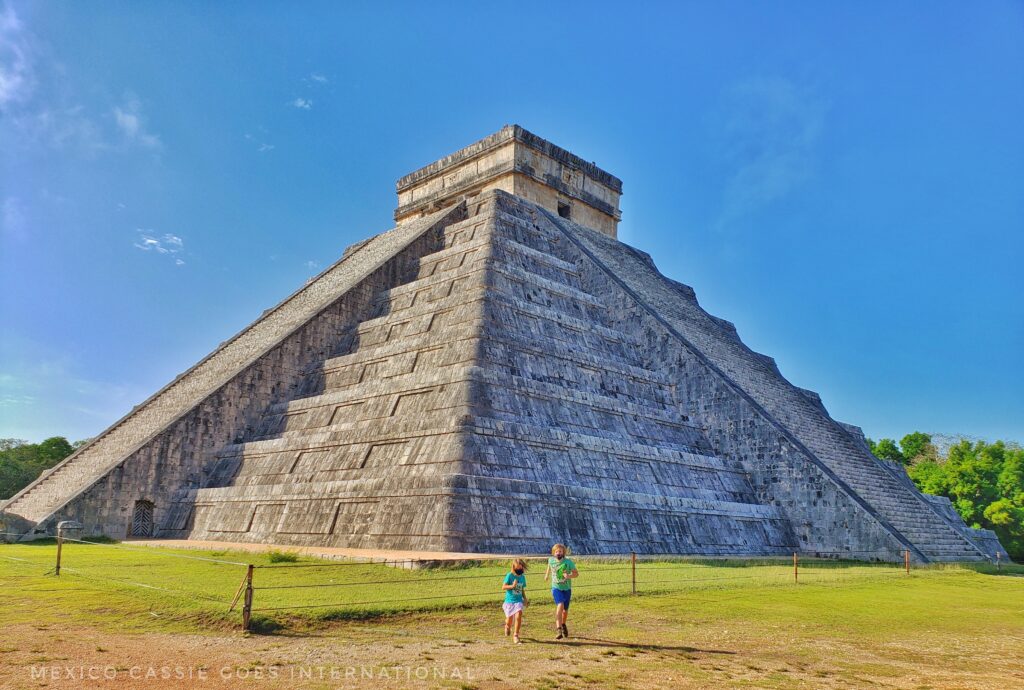 Chichen Itza pyramid with two small children running away from it, towards camera. blue sky