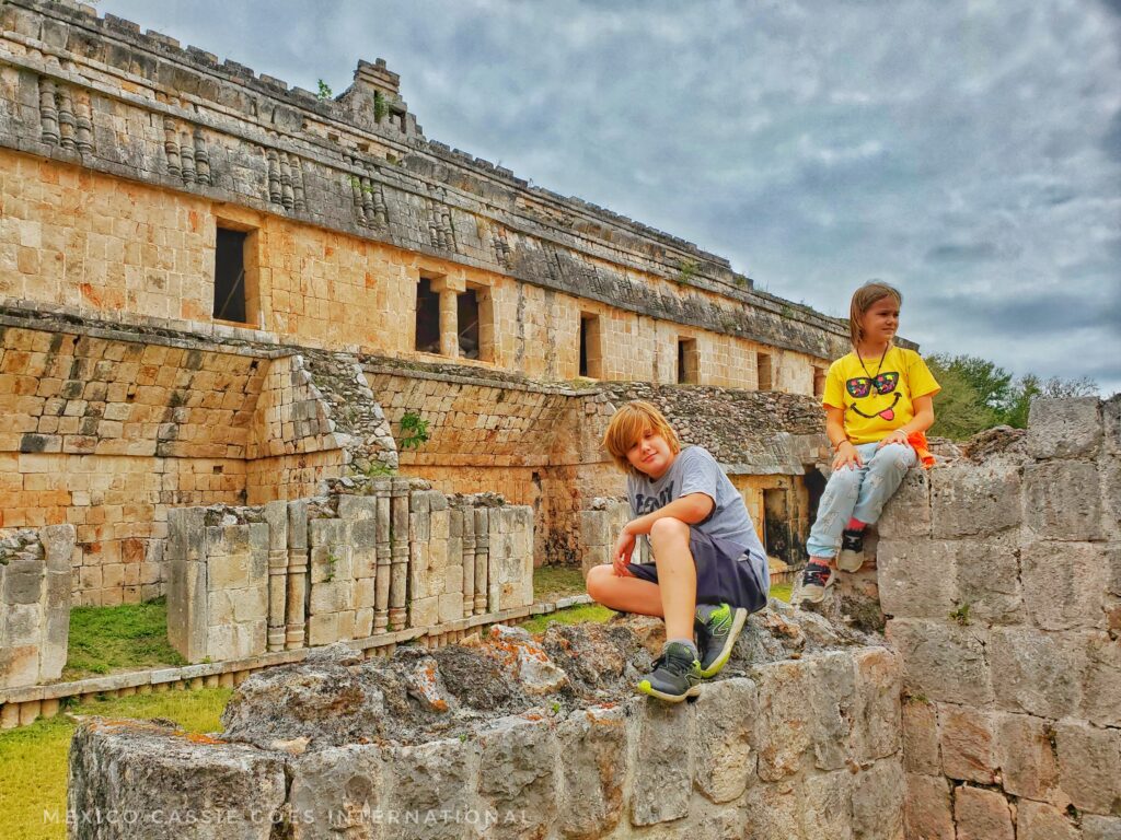 deux enfants assis sur un mur maya avec un long bâtiment maya derrière eux