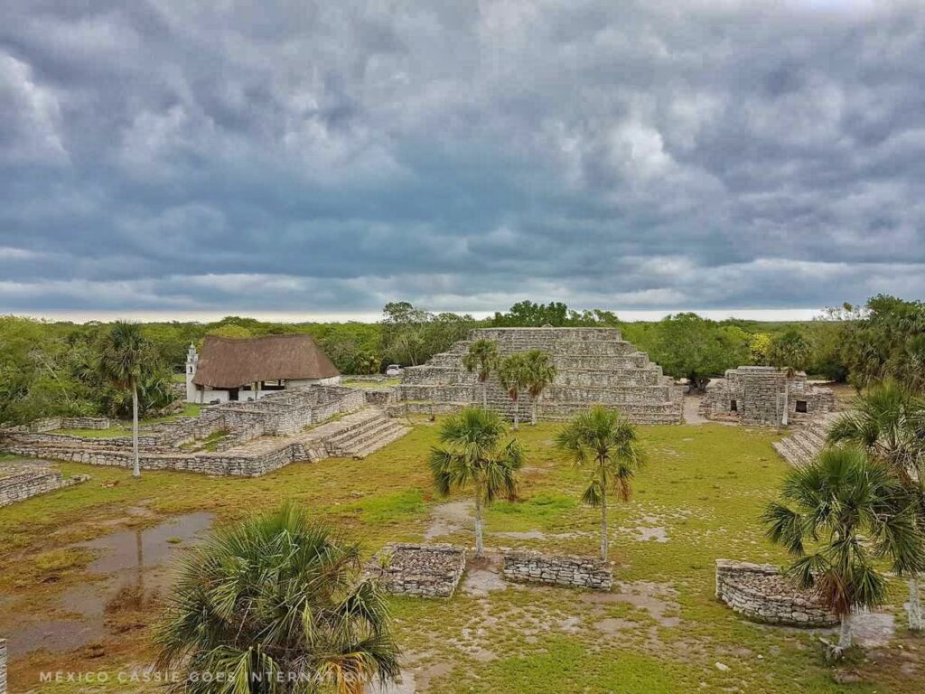view over xcambo ruins, pyramid in front, thatched chapel to left. ruined outlines on floor and palm trees. heavy, grey sky
