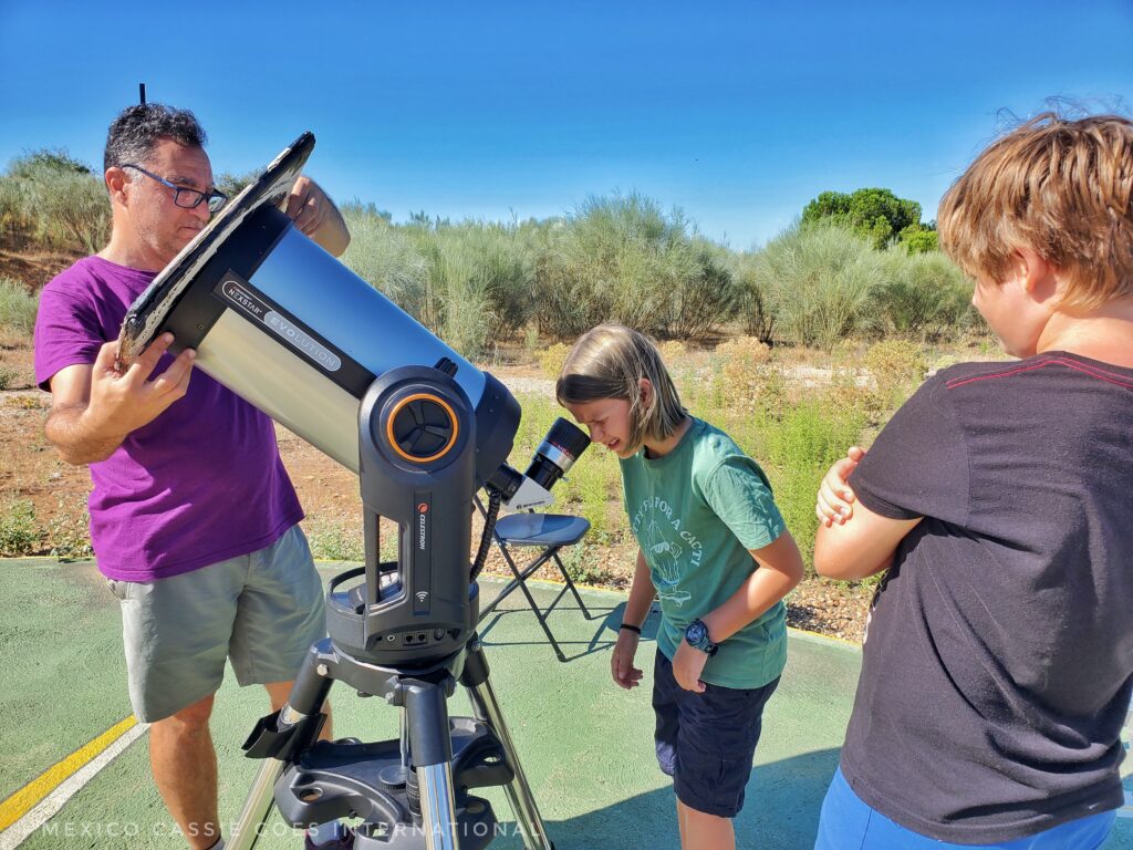 child in green tshirt looking into large telescope. man in red shirt holds protector over lens (daylight) 