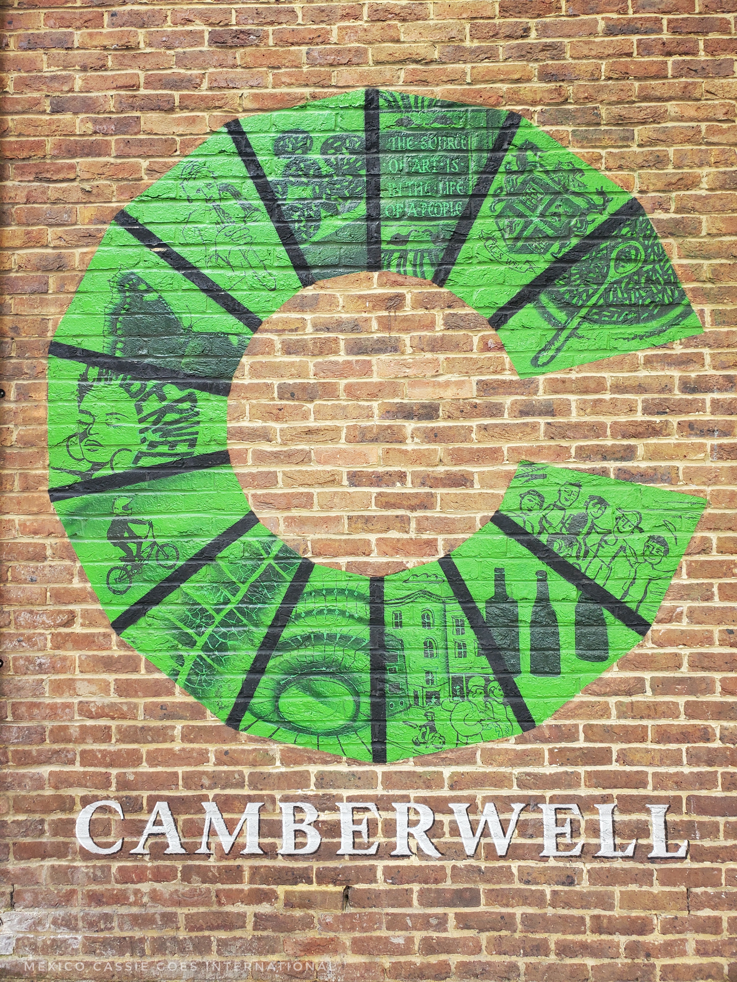 Green C painted on brick wall. Camberwell written underneath in white capitals. C is split into13 sections each with its own picture of something to do with Camberwell