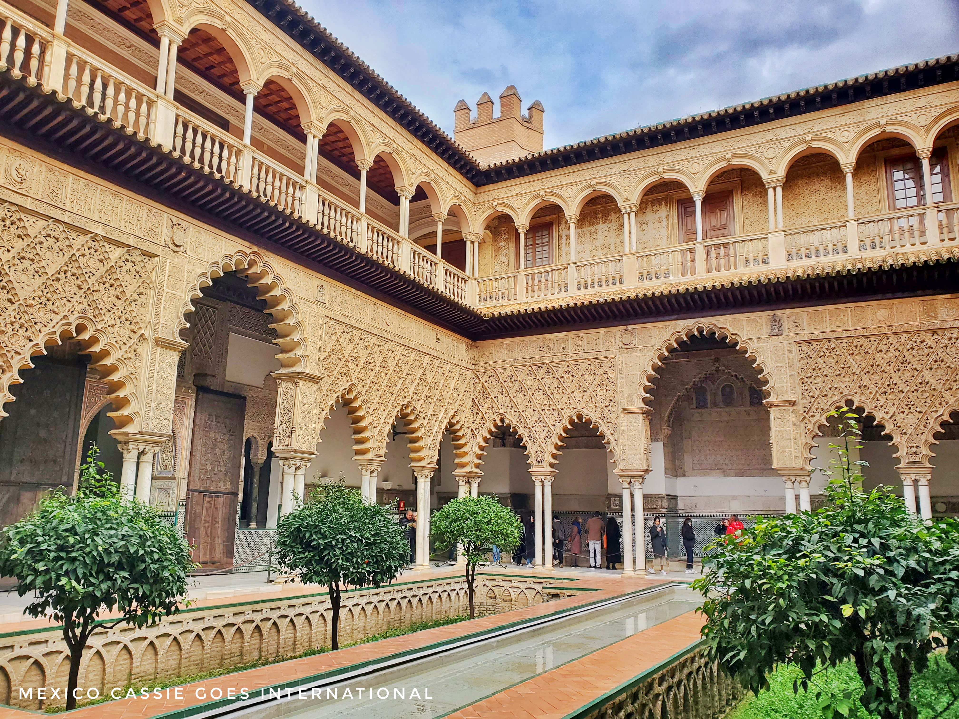 view of the main patio in the alcazar of seville. reflecting pool, small trees and 2 storey building with many arches