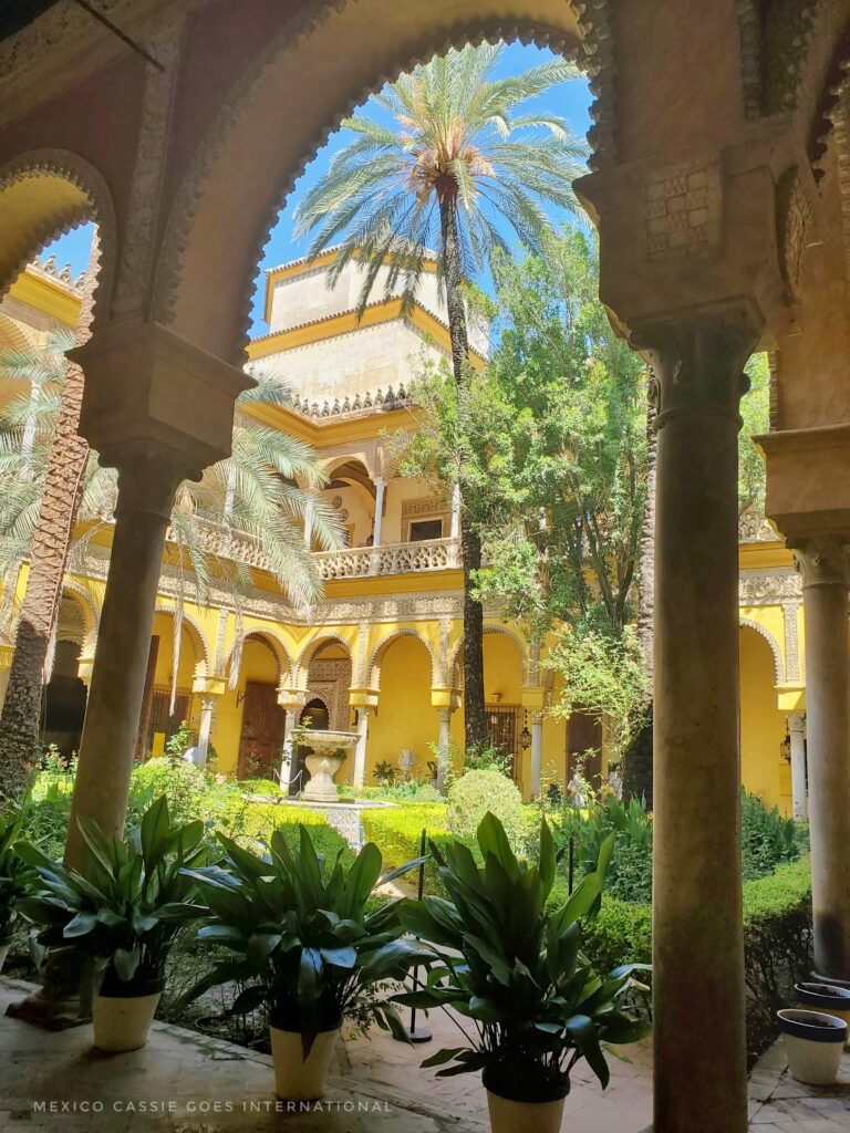 Moorish courtyard viewed through arch. Yellow building, green trees, one large palm tree