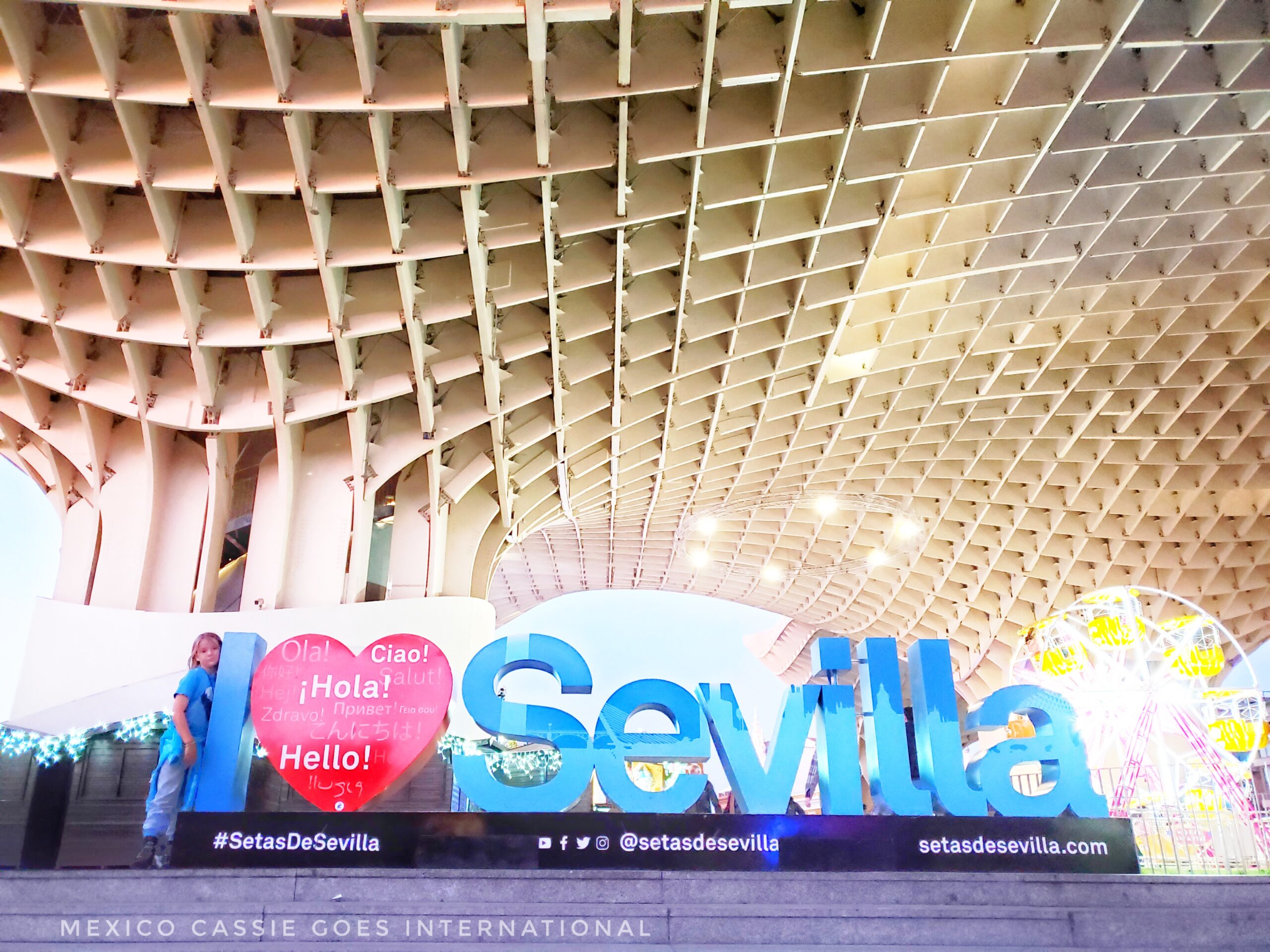 I ❤️ Sevilla in large letters with kid standing next to the I. Wooden criss cross sculpture in background