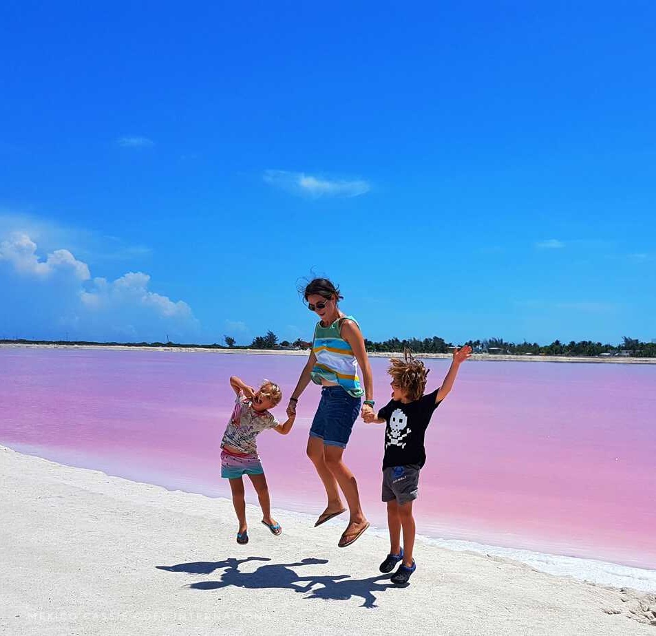 adult and two kids jumping while holding hands in front of pink water. bright blue sky.