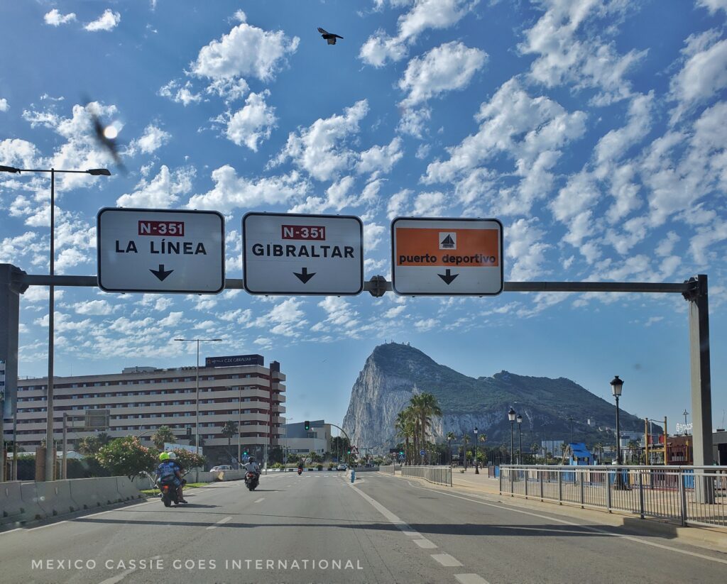 road leading to gibraltar: rock of gibraltar in distance. three signs over road: la linea, gibraltar and puerto deportivo