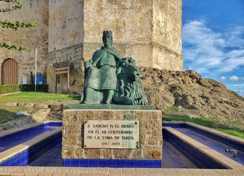 statue of sancho el bravo sitting with a lion at base of castle. plaque reads: "a sancho IV el bravo en el VII centenario de la toma de Tarifa 1292 - 1992)