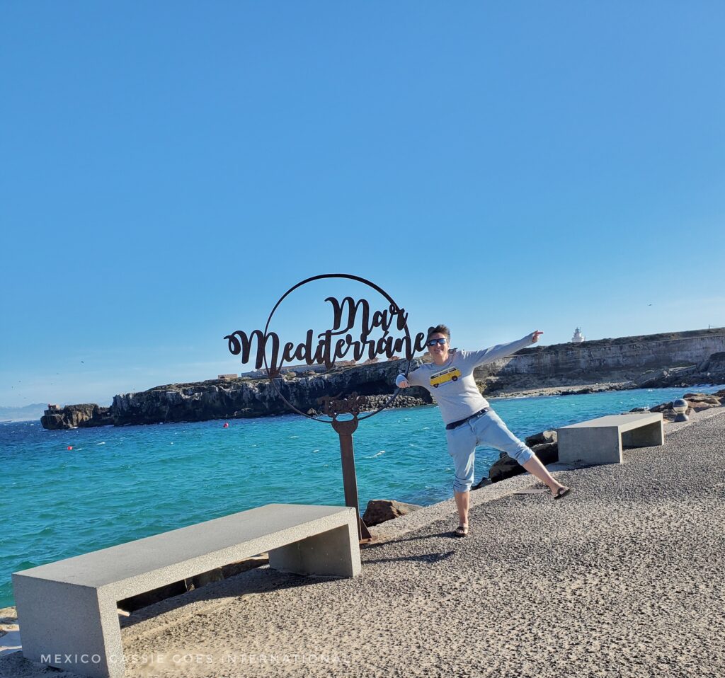 Cassie standing on one leg while holding onto a circular signpost that says "Mar Mediterraneo in front of the sea