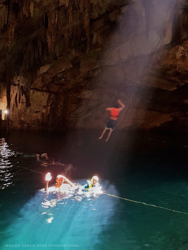 man and child in water with sunlight bathing them. person in air above them having just let go of a rope swing. all in a cave