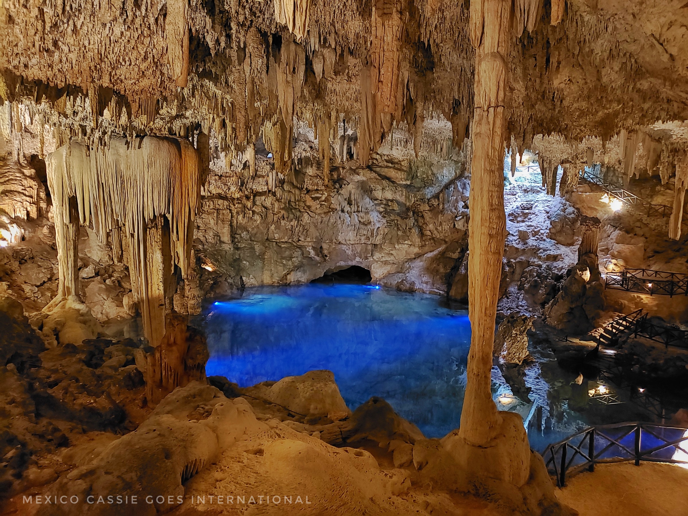 bright blue water at bottom of cave. cave is full of stalactites and stalagmites