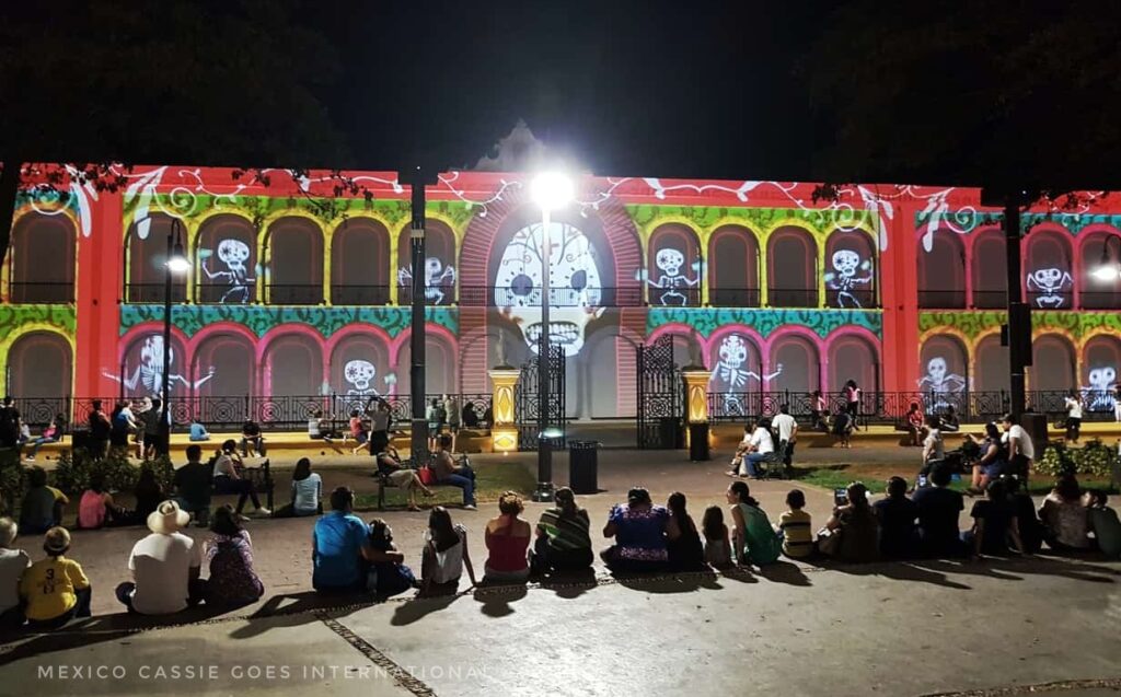 people sitting on paved floor watching pictures mapped onto building in front. picture is off a white skull in middle and dancing skeletons in the arches of the building are highlighted