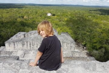 child in black sitting on top of stone ruin looking back at camera - more ruins and jungle in front of him