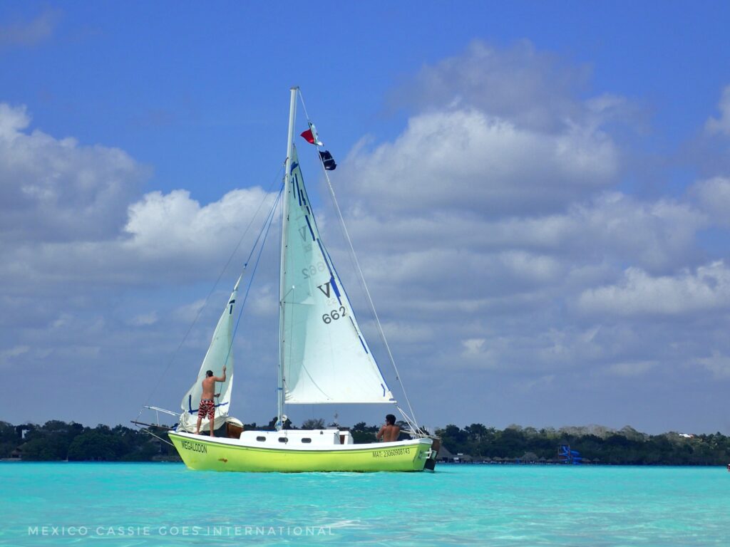 yellow boat with white sails on stunning turquoise blue water. two men on boat, one sitting, one hoisting a sail