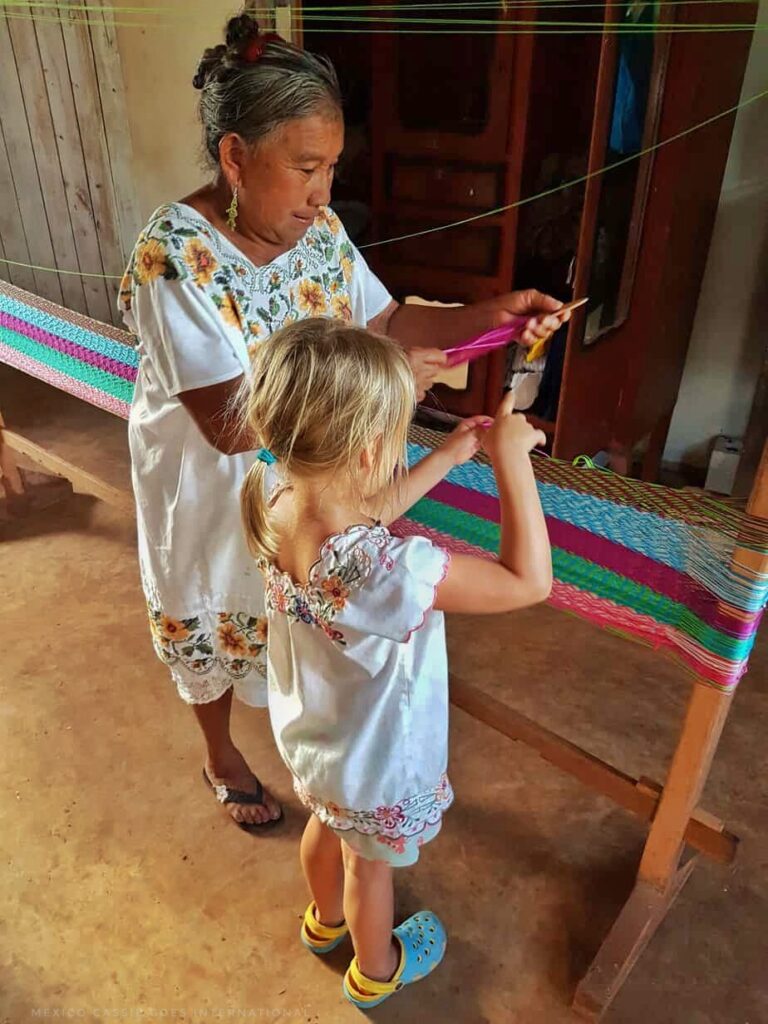 old Maya woman in traditional white dress with flowers teaching young blond girl in traditional white dress how to make a hammock