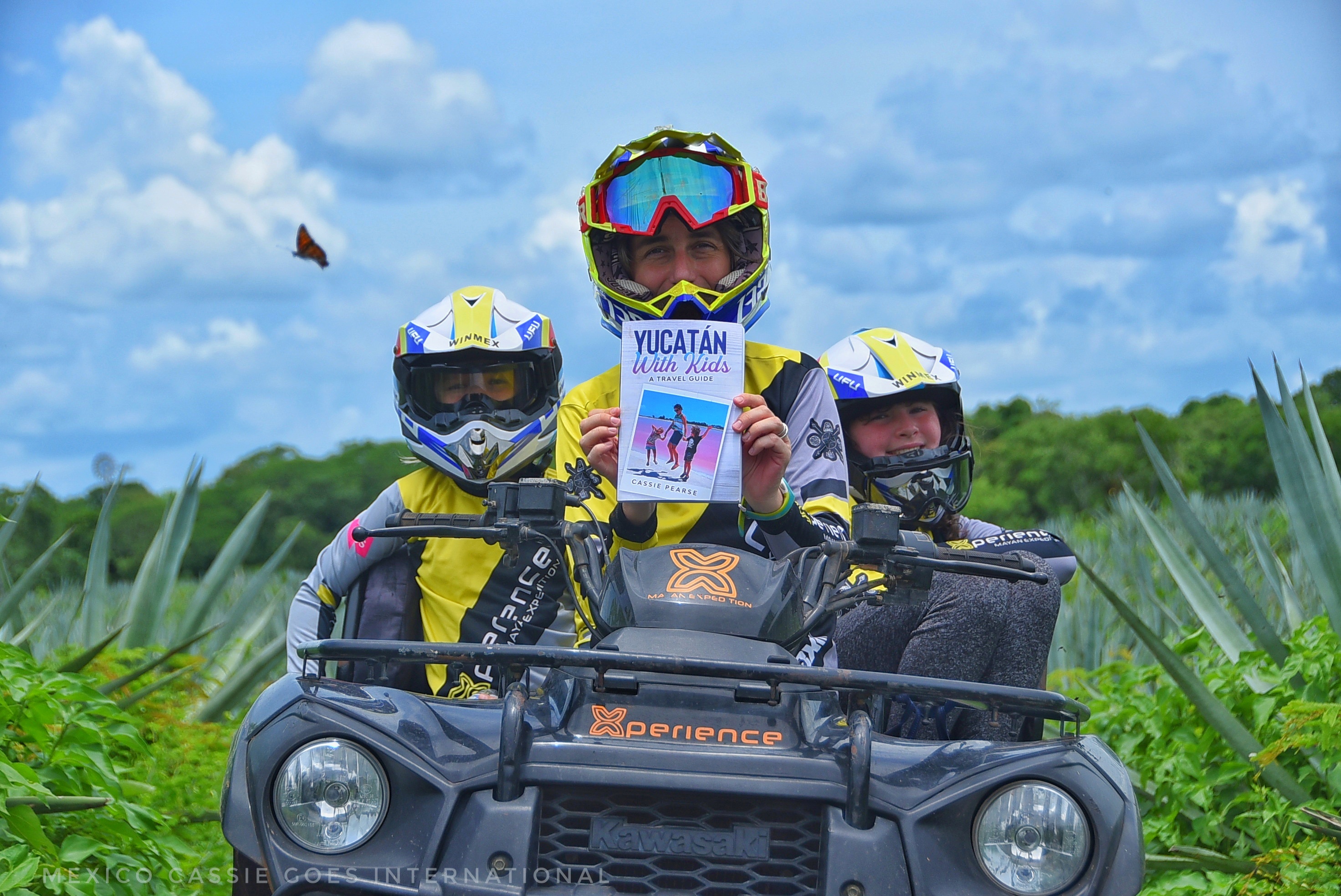 Adult and two children on an ATV. All with helmets and yellow/grey tops. Adult holding a copy of Mexico Cassie's Travel Guide to Yucatan