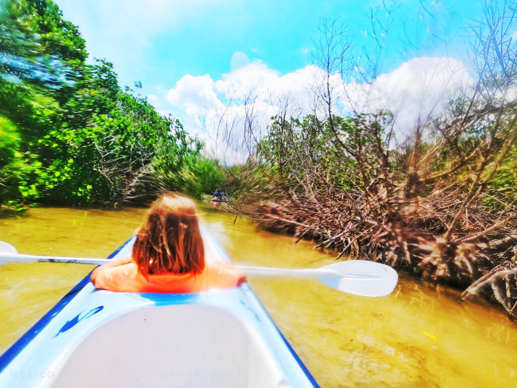 girl in orange tshirt in front of kayak facing away from camera, paddle across lap. Brownish water and green bushes