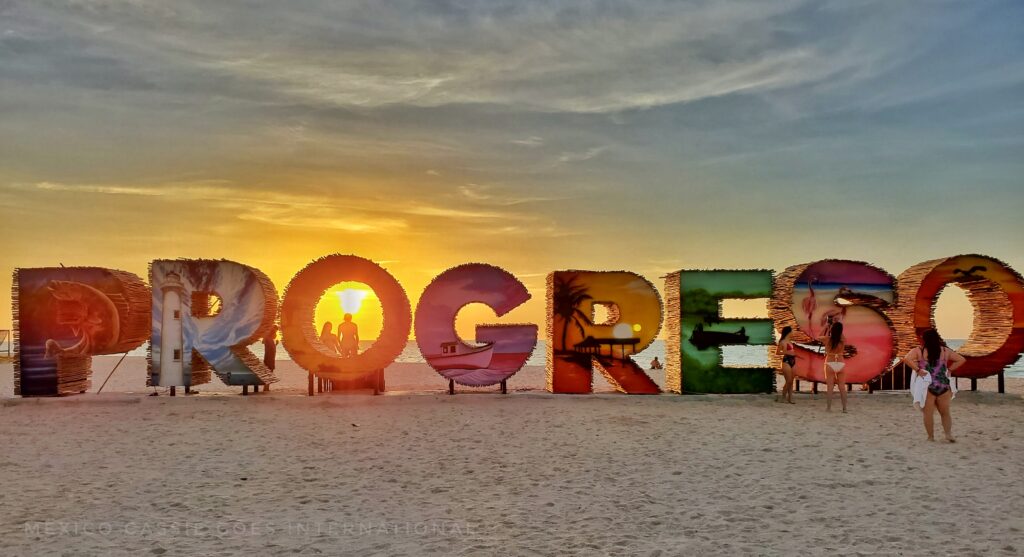 Progreso written in large letters on beach. people milling around the letters. Setting sun shining through the first O