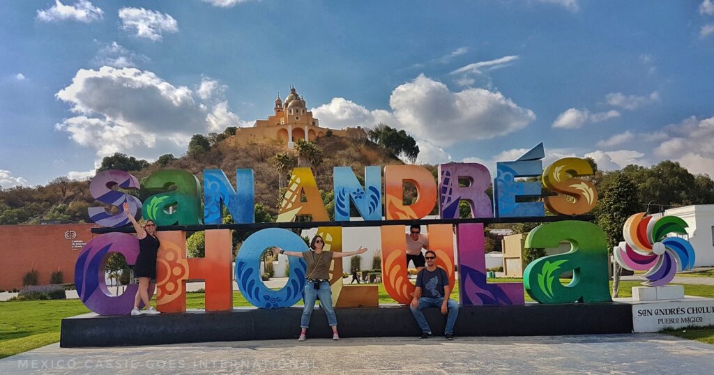 Colourful letters reading San Andres Cholula - four adults standing by the letters. Large yellow church on hill above
