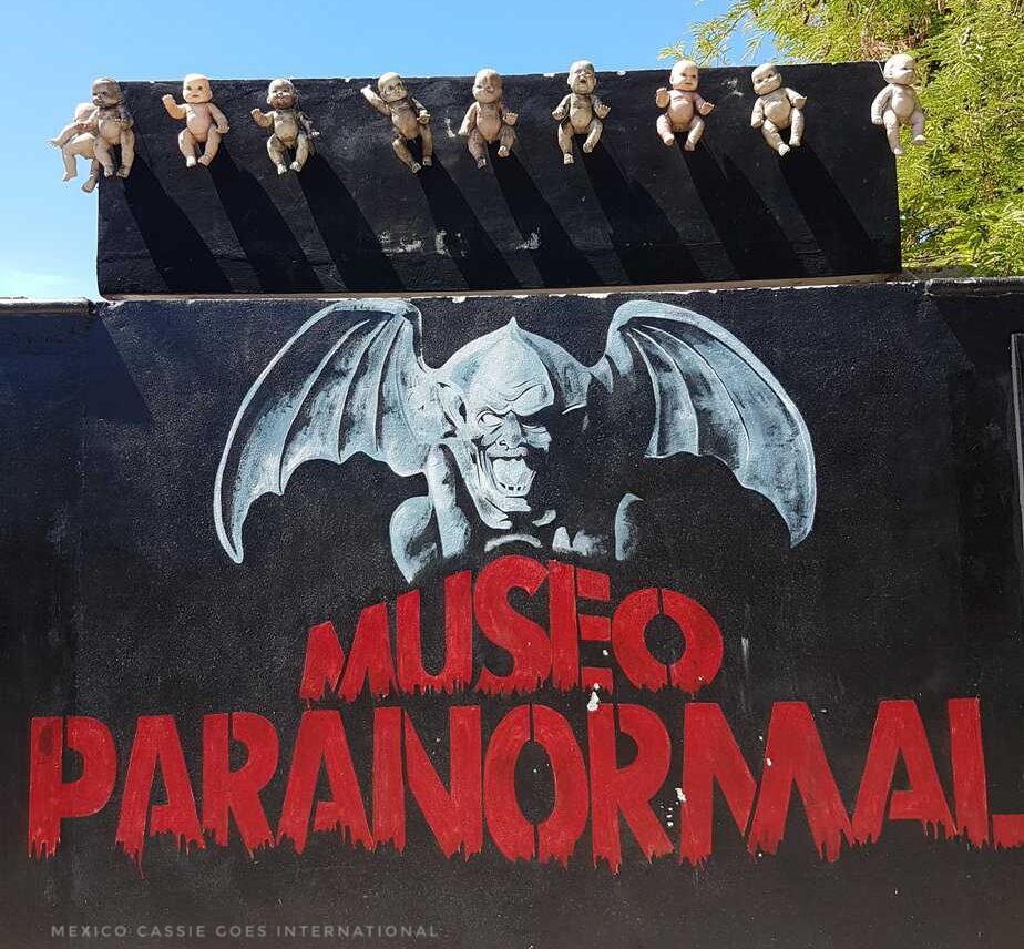 wall painted black with words "museo paranormal" in red, gargoyle painted above. naked dolls hanging above that.