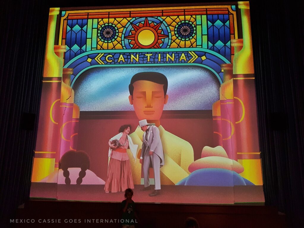 picture of a stylised stage - cantina written below fake stain glass window. man in white on backdrop of stage and man and woman in front. 