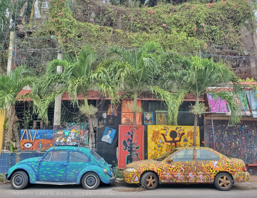 two cars facing left. A beetle painted blue and a second car painted yellow. Both with patterns on top of paint. Trees and buildings behind