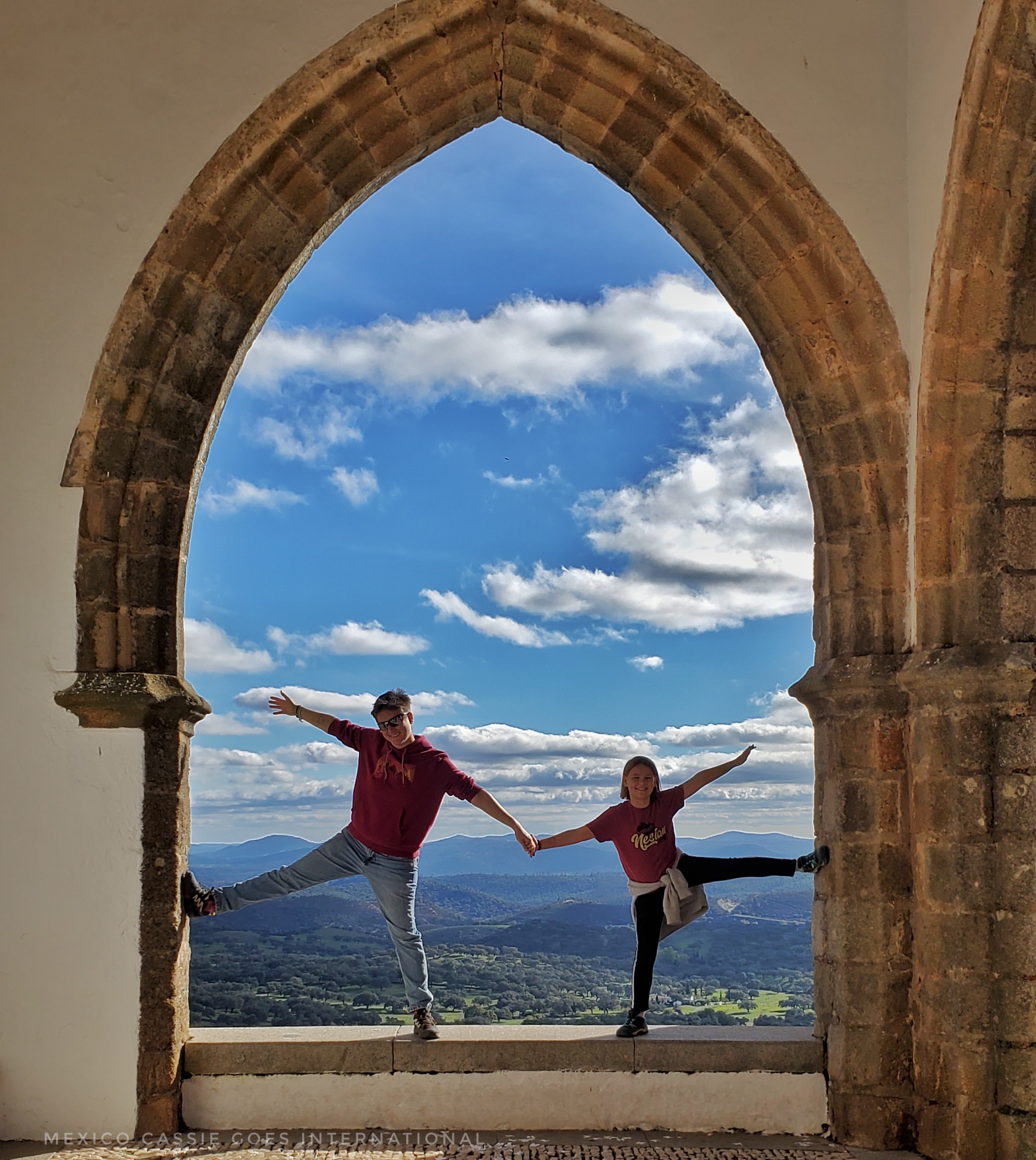 adult and kid standing in large vaulted window both on one leg with other leg against a wall. They're holding hands.