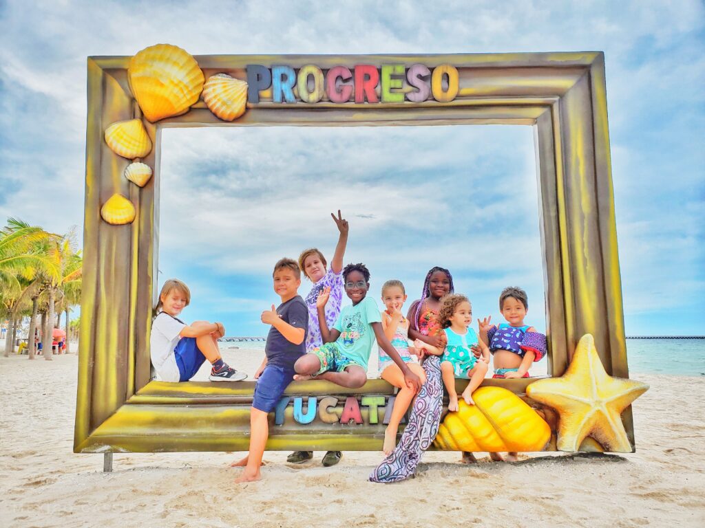 group of children sitting in a large photo frame on a beach. Says Progreso at top