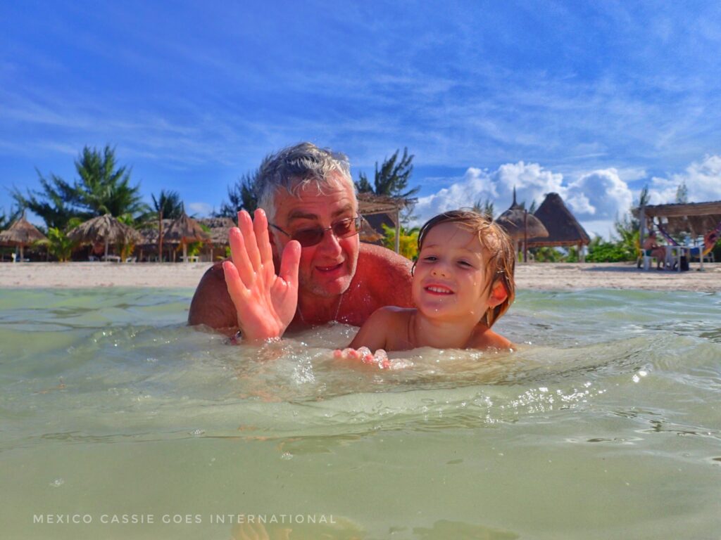 man and small child in very shallow sea water. both looking at camera, man waving. palms and blue sky behind them