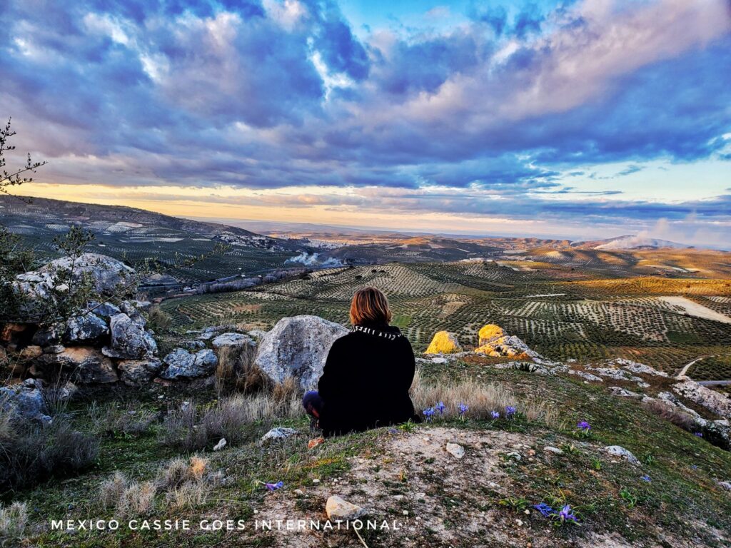 person in black sitting on rock looking out at expanse of fields under a setting sun