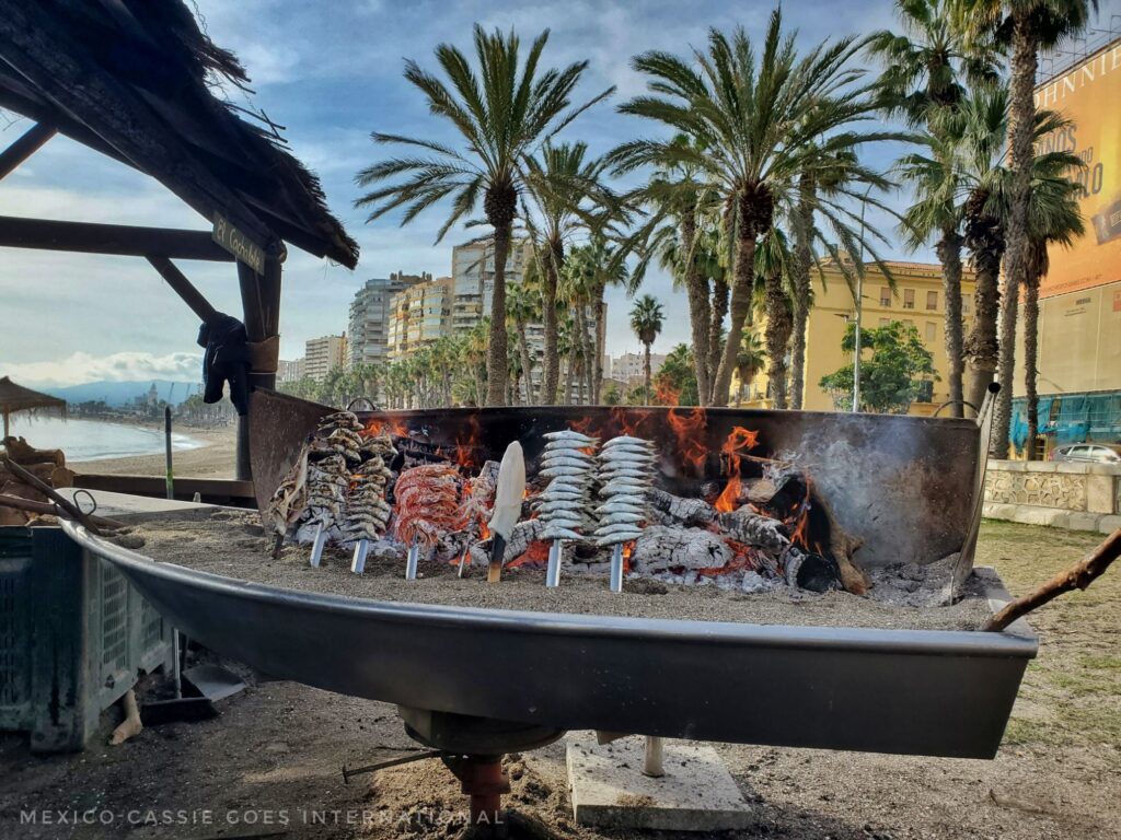 sardines on skewers cooking on a boat shaped bbq. palm trees in background