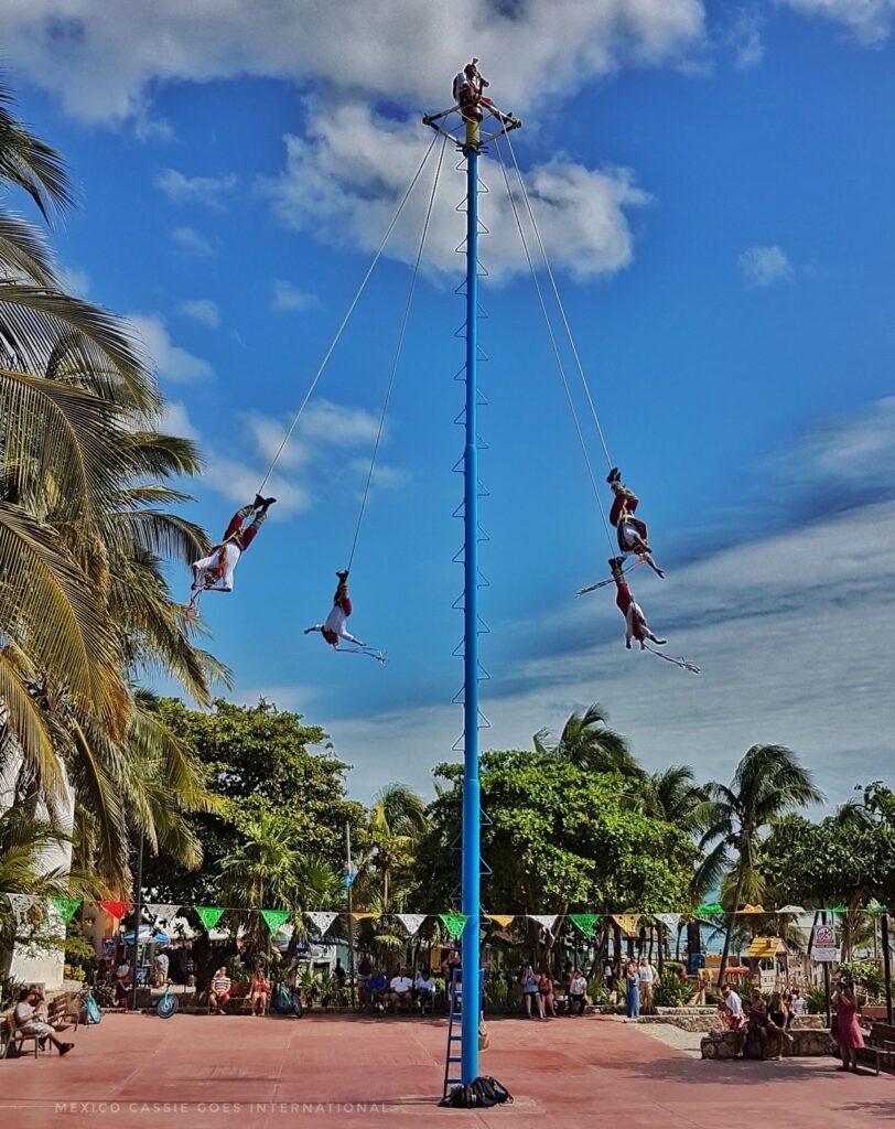 blue sky, palm trees, tall blue pole with four men hanging upside down (dancers) from it