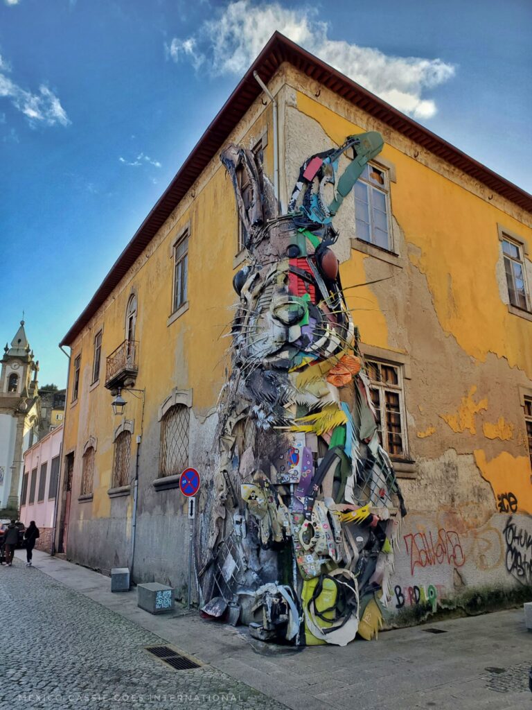 Photo of yellow two storey house. Giant mural of a rabbit over one corner- one side made of brightly coloured trash, other side of grey trash