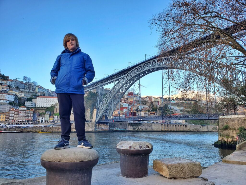 boy in blue jacket standing in  front of river with an iron bridge behind him