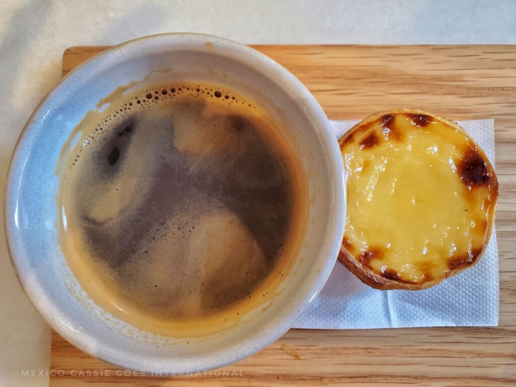 view of a cup of black coffee in a white mug sitting next to a pastel de nata on a white napkin. Both on a wooden board