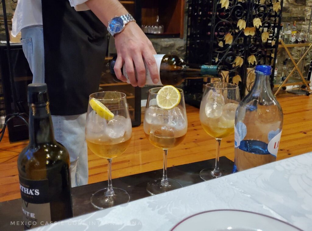 hand wearing big silver watch pouring white port into wine glasses that contain ice and lemon, bottle of port on left and water on right