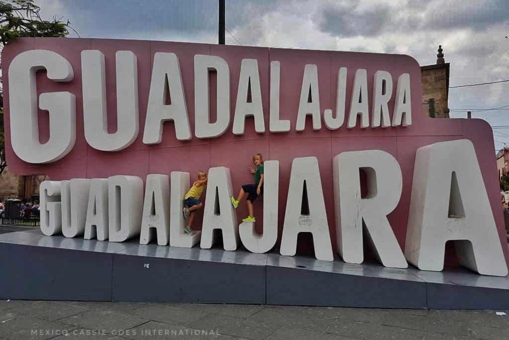 guadalajara written twice, once above the other in big white letters on red background. 2 small kids sitting on either side of middle "A" of bottom text