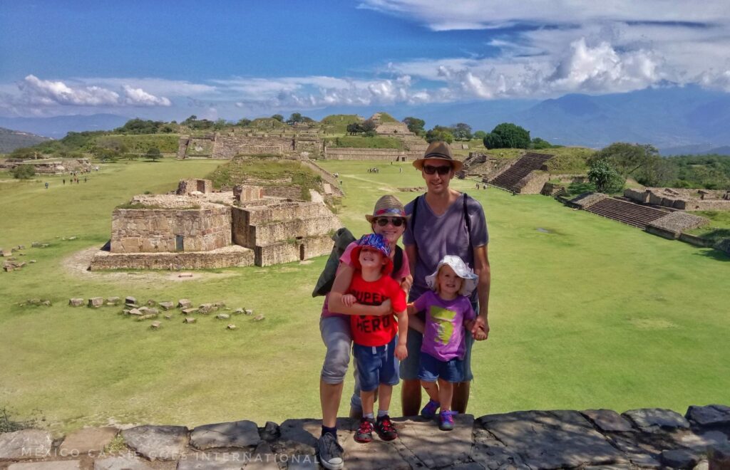 family (2 adults and 2 small kids) standing on steps above grass and ruins of monte alban