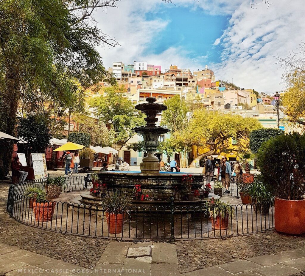 plaza with fountain surrounded by small railing, buildings behind rising on a hillside