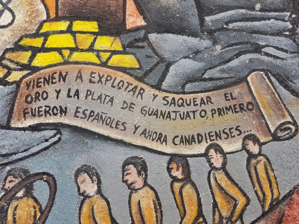 section of a mural - grey colouring, workers in yellow, bars of gold, scroll reading vienen a explotar y saquear el oro y la plata de guanajuato primaero venieron españoles y ahora canadienses