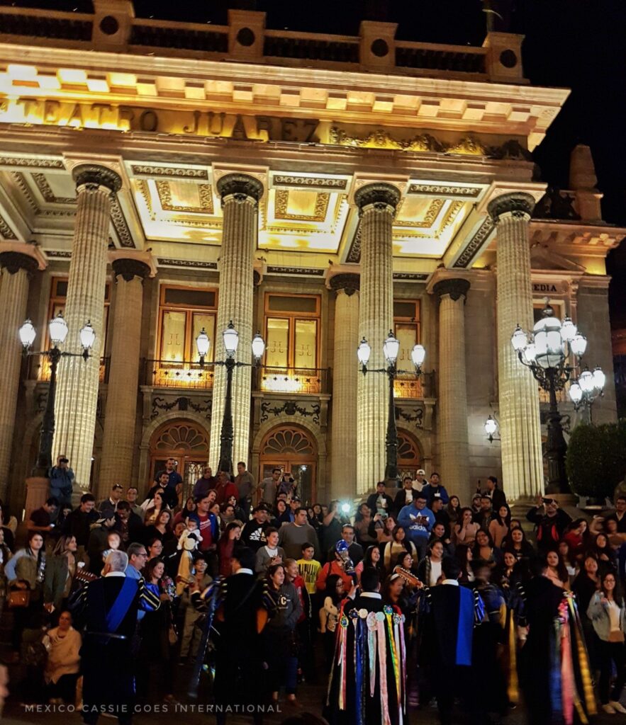 theatre steps at night crowded with people, the people in the front are in long black gowns with coloured strips hanging down