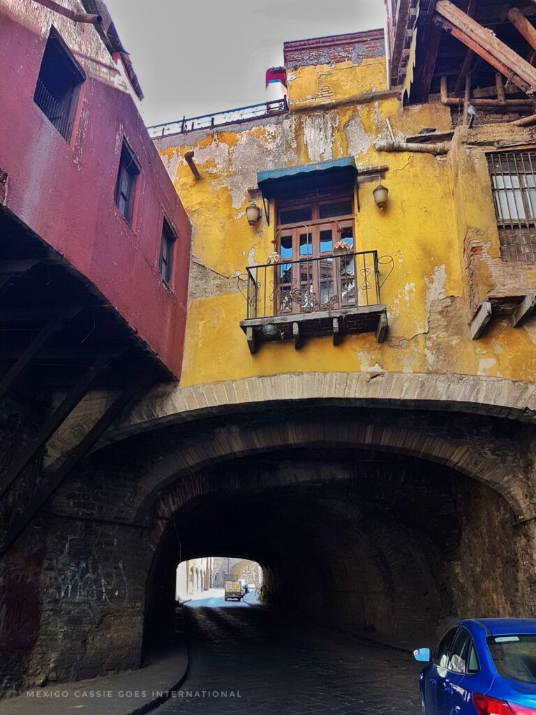 dirty yellow house over an arch tunnel road. red building to left. blue car on right