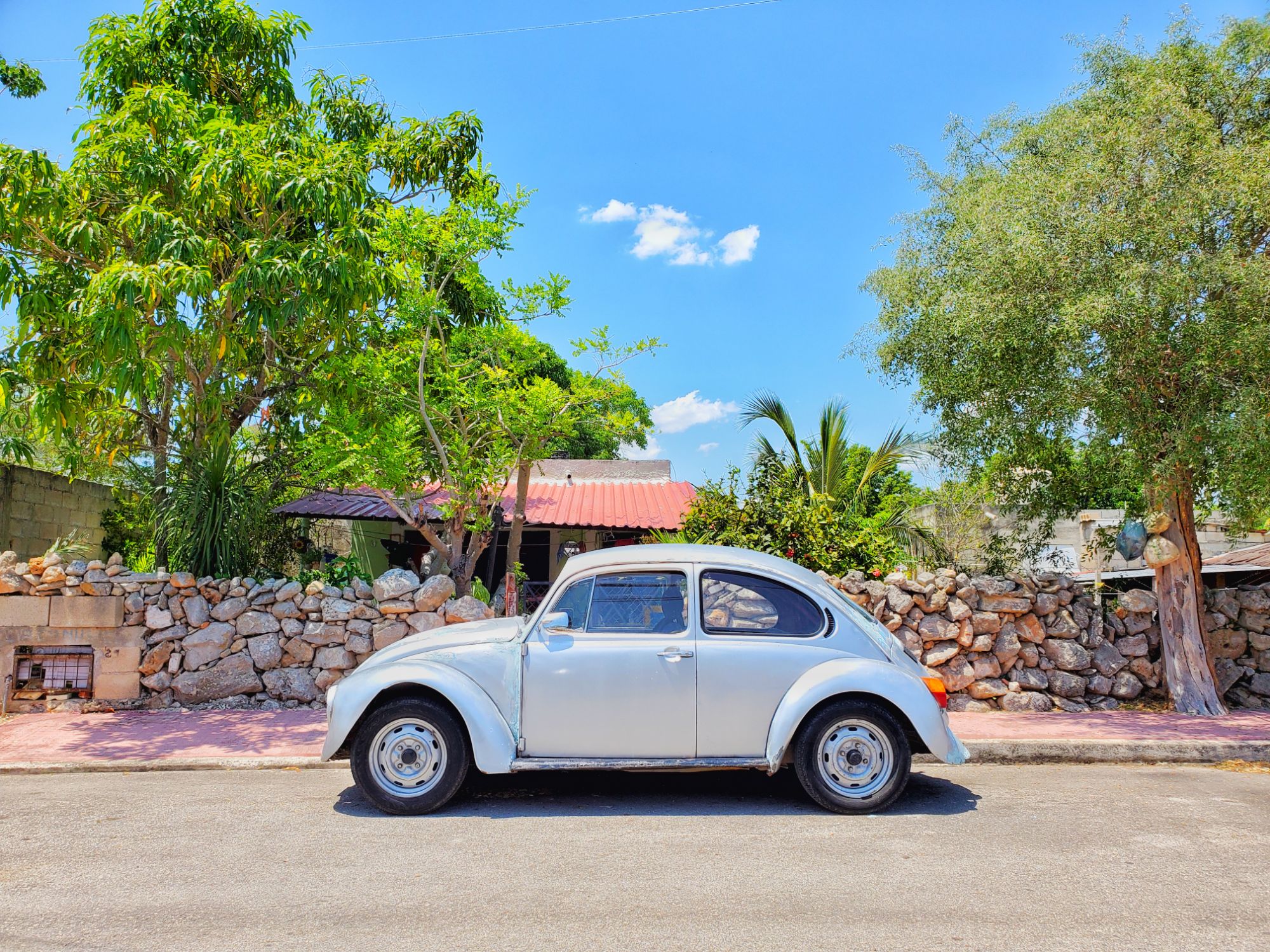 white vw in front of a stone wall, trees, red roof house and blue sky
