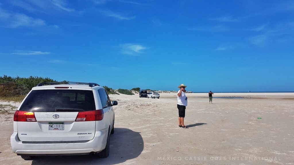 white car on beach, man in white tshirt lookig at camera (from afar)