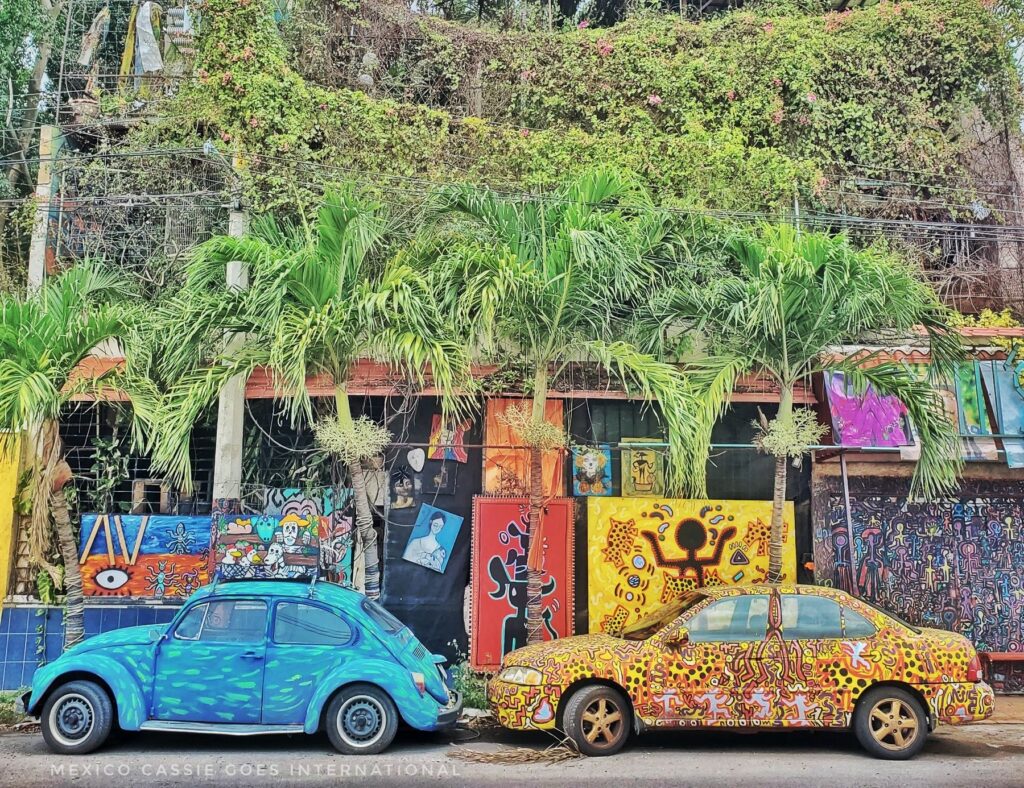 two parked cars in front of a brightly painted building. car on left is a bright blue decorated VW, on right is decorated like a jaguar
