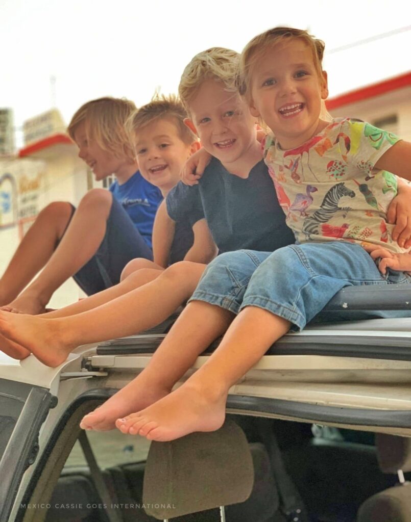 four small kids sitting on roof of car - all smiling, 3 looking at camera